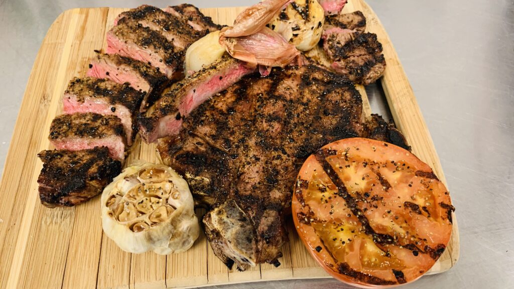 Grilled steak with roasted garlic and grilled tomato served on a wooden board.