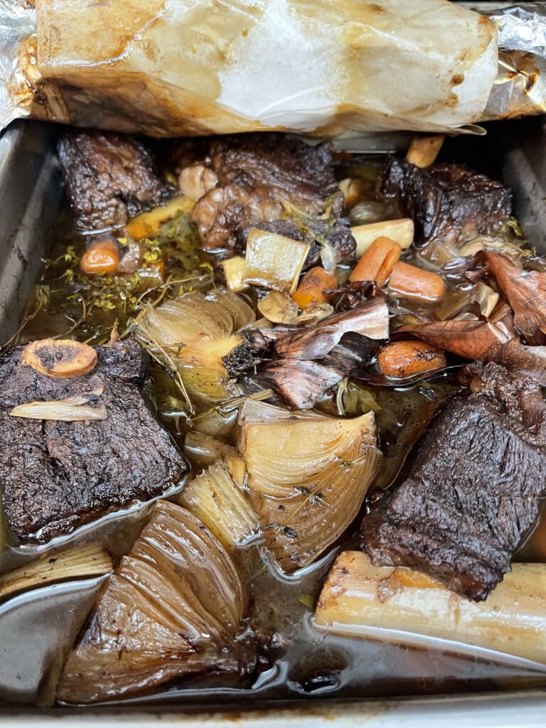 Alt Text: Beef short ribs braising in red wine, beef stock, onions, carrots, celery, garlic, and fresh thyme before a long slow cook.