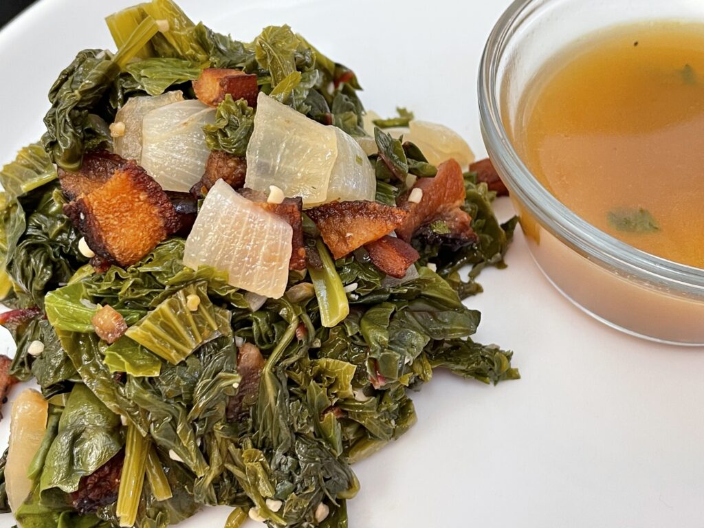 A plate of Southern mixed greens made with red chard, spinach, and mustard greens, topped with smoked bacon and sweet onions, served with a side of flavorful pot liquor.